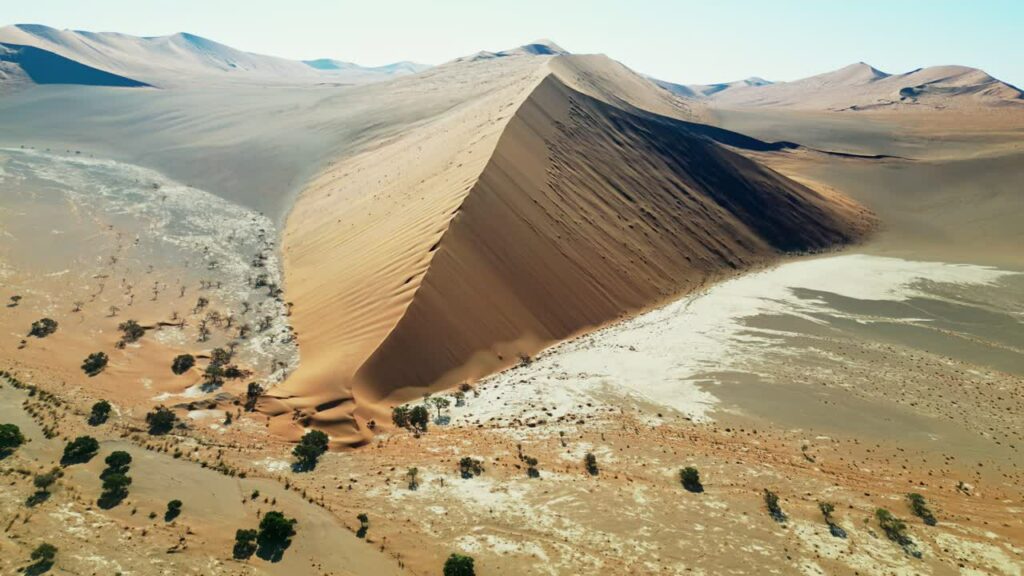 Video - Aerial drone view of a massive triangular sand dune rising above the desert plains in Namibia with sparse desert vegetation