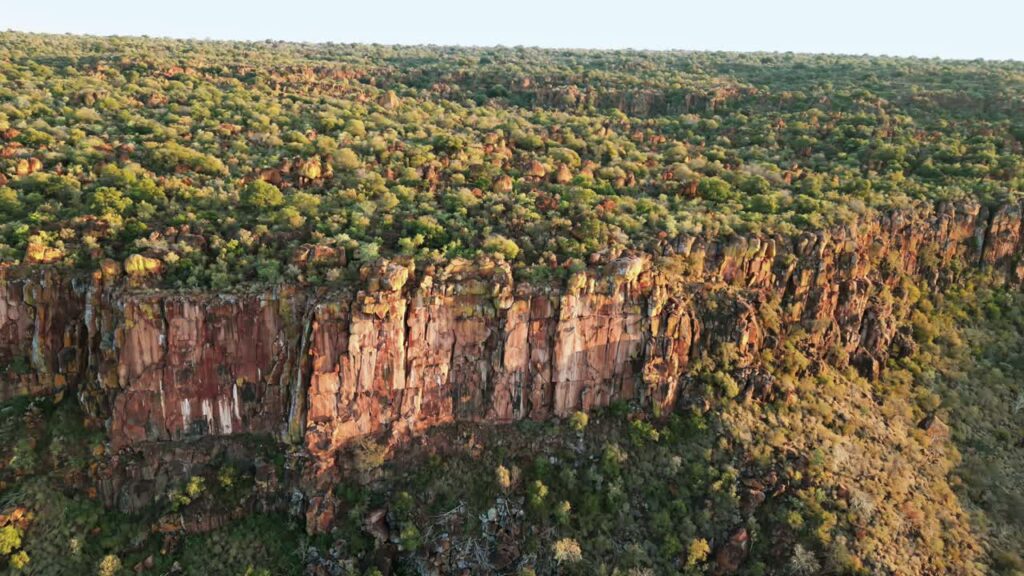 Video - Aerial drone view of dramatic rocky cliffs rising above a green savanna landscape in Namibia during warm golden hour light