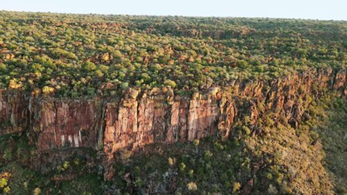 Video - Aerial drone view of dramatic rocky cliffs rising above a green savanna landscape in Namibia during warm golden hour light