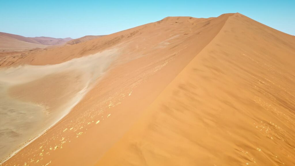 Video - Aerial drone view along the ridge of a massive sand dune in the Namib Desert with smooth wind shaped patterns and golden desert landscape