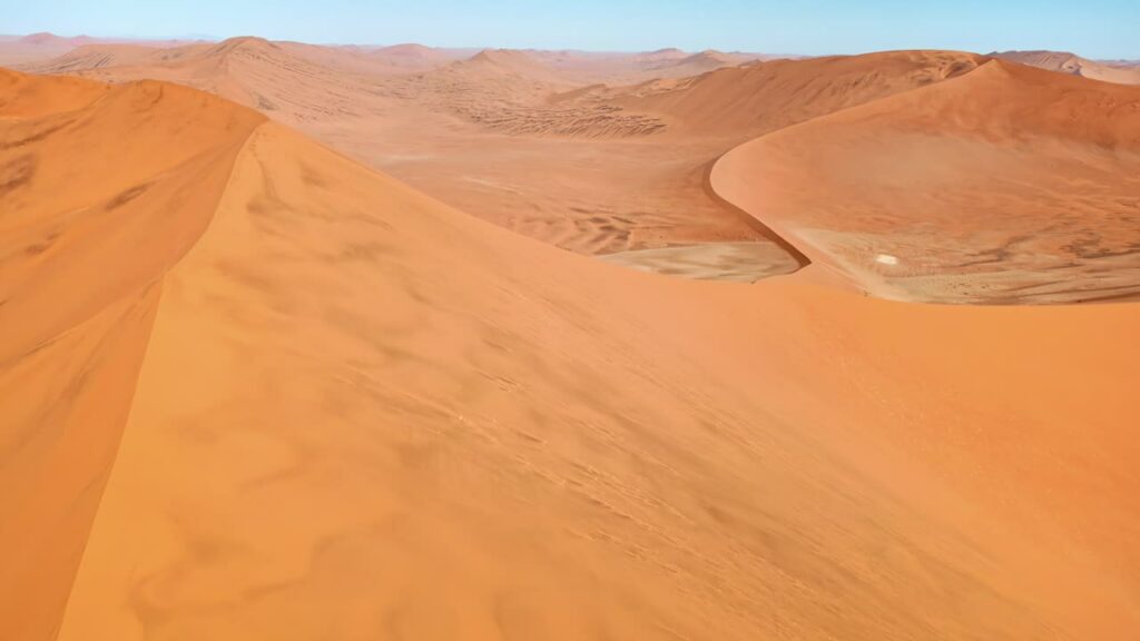 Video - Aerial drone view along the ridge of a massive sand dune in the Namib Desert with smooth wind shaped patterns and golden desert landscape