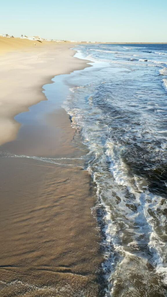 Video - Aerial drone view of the remote Skeleton Coast shoreline in Namibia showing waves breaking along a vast sandy beach and desert dunes. Vertical