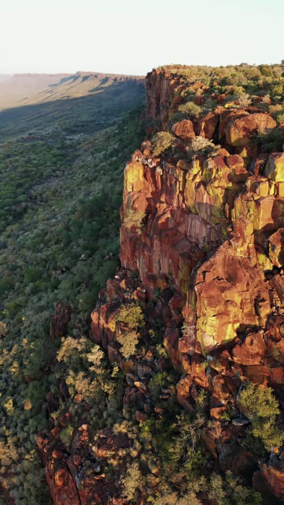 Video - Aerial drone view of dramatic rocky cliffs and green vegetation in Namibia during warm sunset light overlooking a vast wilderness valley. Vertical