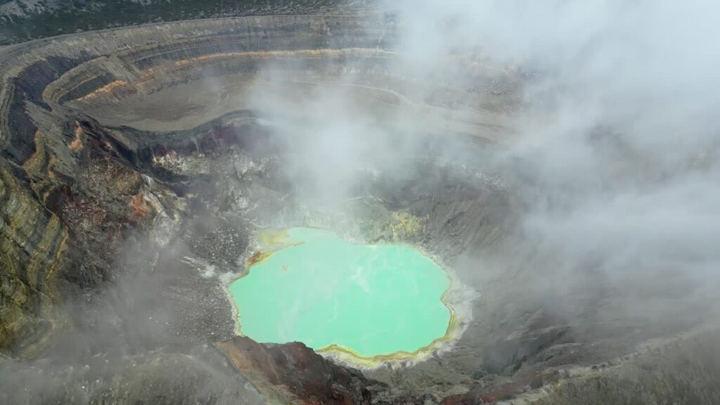 Video - Aerial drone view of a turquoise crater lake inside a volcanic crater in Guatemala, surrounded by rugged volcanic rock formations and geothermal landscape