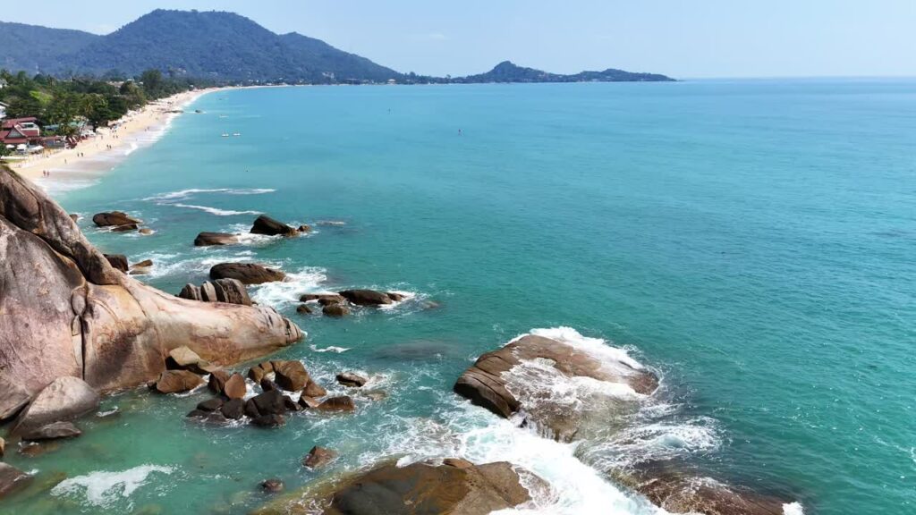 Video - Aerial drone view of ocean waves breaking against large coastal rocks in turquoise water in Thailand