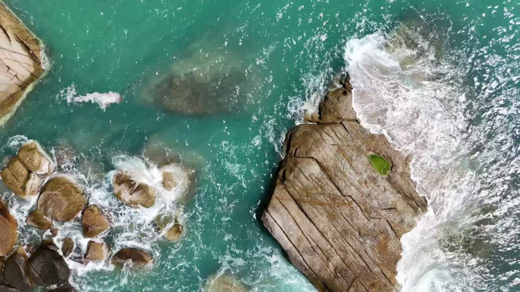 Video - Aerial drone view of ocean waves breaking against large coastal rocks in turquoise water in Thailand