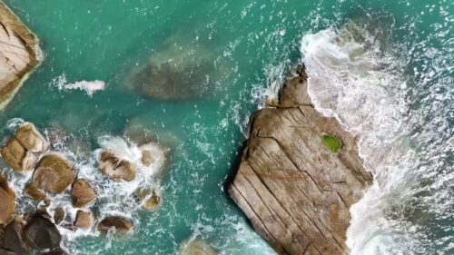 Video - Aerial drone view of ocean waves breaking against large coastal rocks in turquoise water in Thailand