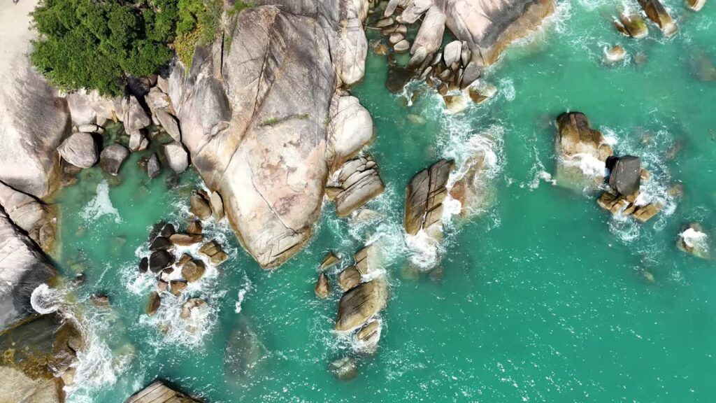 Video - Aerial drone view of ocean waves breaking against large coastal rocks in turquoise water in Thailand