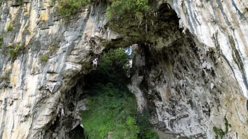 Video - Aerial drone view of a large cave entrance inside a dramatic limestone cliff surrounded by dense tropical vegetation