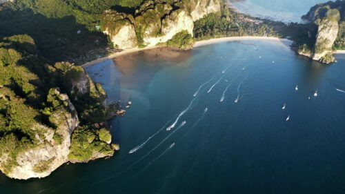 Video - Aerial drone view of boats moving across turquoise water near dramatic limestone cliffs and beach in Krabi, Thailand