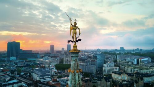 Video - Aerial drone cinematic view of statue of Saint Michael atop the spire of the Brussels Town Hall at sunset in Belgium. Multiple buildings on the background