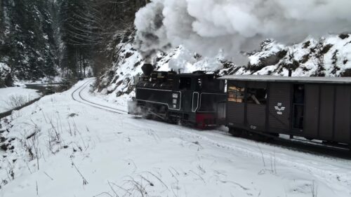 Video - Aerial drone view of the moving steam train Mocanita in a valley along a river in winter, hills covered with bare forest and snow on mountains