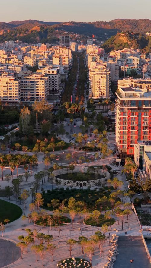 Video - Aerial drone view of the golden hour lighting highlighting Vlora's central urban park, featuring circular pathways, palm trees, playgrounds, and landscaped green spaces. Durres, Albania, Vertical